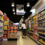 Seorang pembeli mendorong keranjang belanja di lorong supermarket yang penuh produk